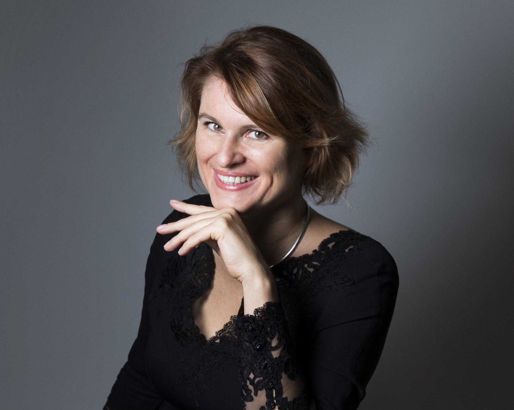 Portrait d’Audrey Alwett souriante, la main sous le menton, sur fond gris